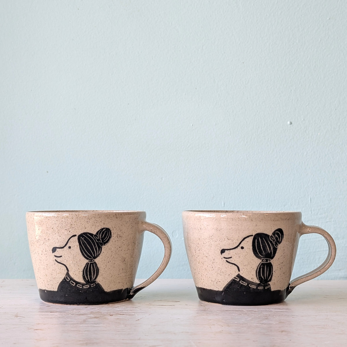 Two handmade speckled beige ceramic mugs with black painted dog illustrations, displayed side-by-side on a light wooden surface against a pale blue wall.