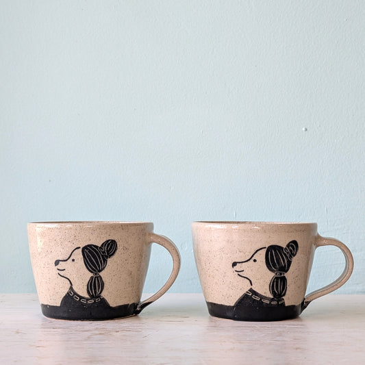 Two handmade speckled beige ceramic mugs with black painted dog illustrations, displayed side-by-side on a light wooden surface against a pale blue wall.
