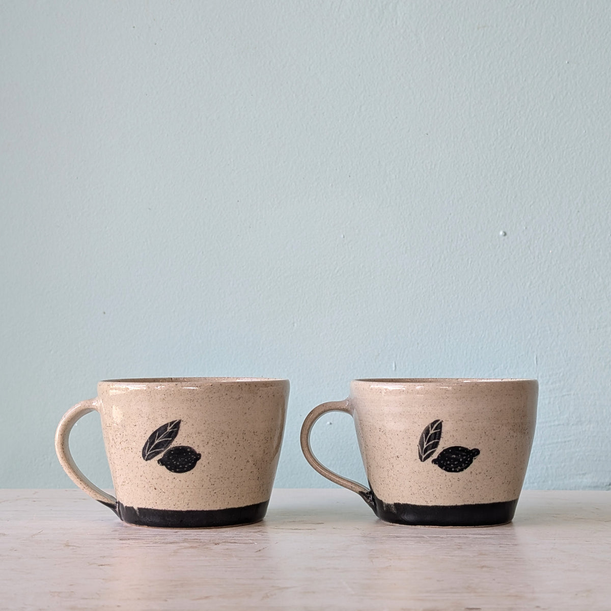 Two handmade ceramic mugs with speckled beige glaze and black leaf motifs, sitting on a light wooden surface against a pale blue wall.