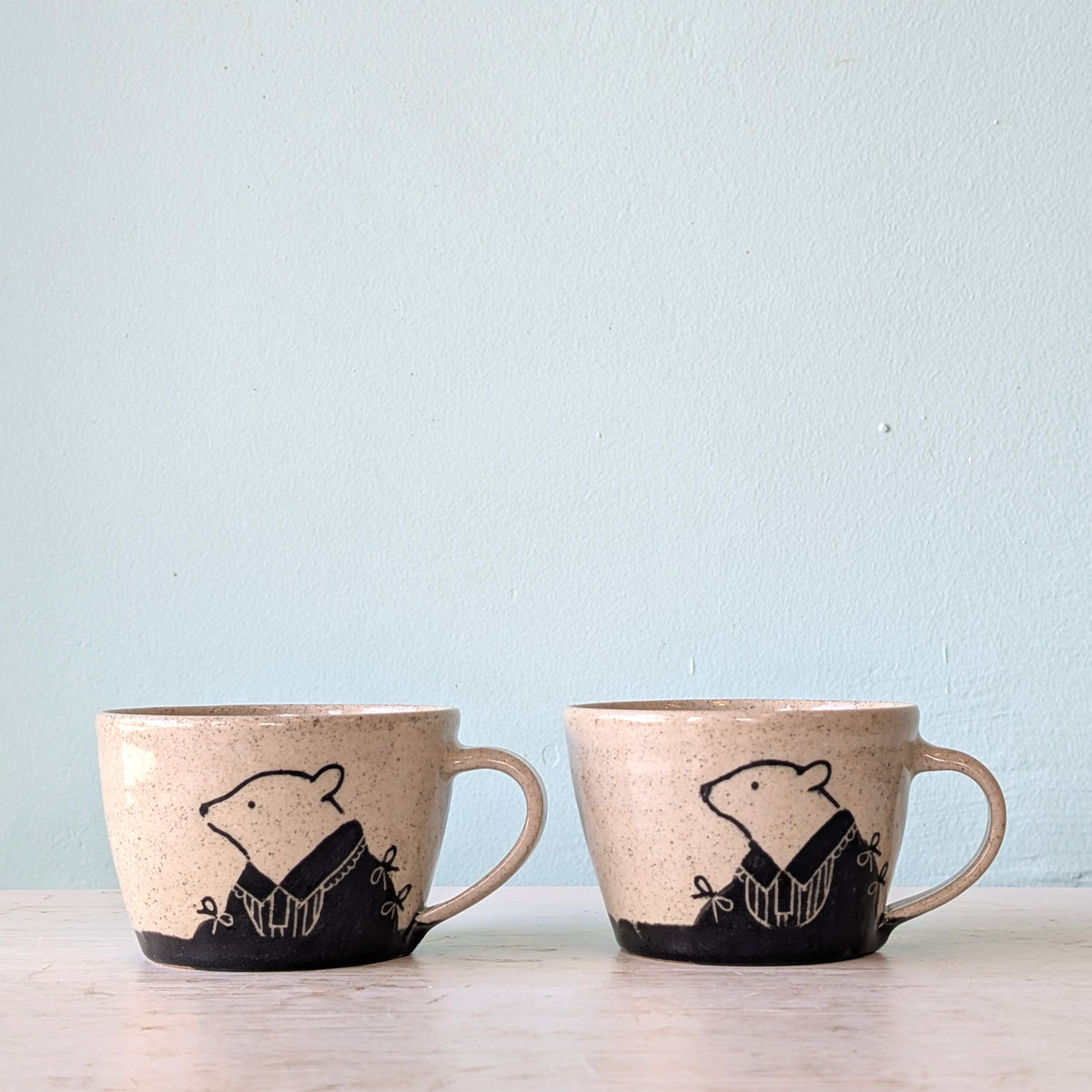 Two speckled beige ceramic mugs with black bear illustrations, displayed side-by-side on a light wooden surface against a pale blue wall.