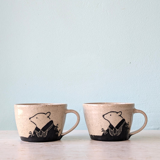 Two speckled beige ceramic mugs with black bear illustrations, displayed side-by-side on a light wooden surface against a pale blue wall.