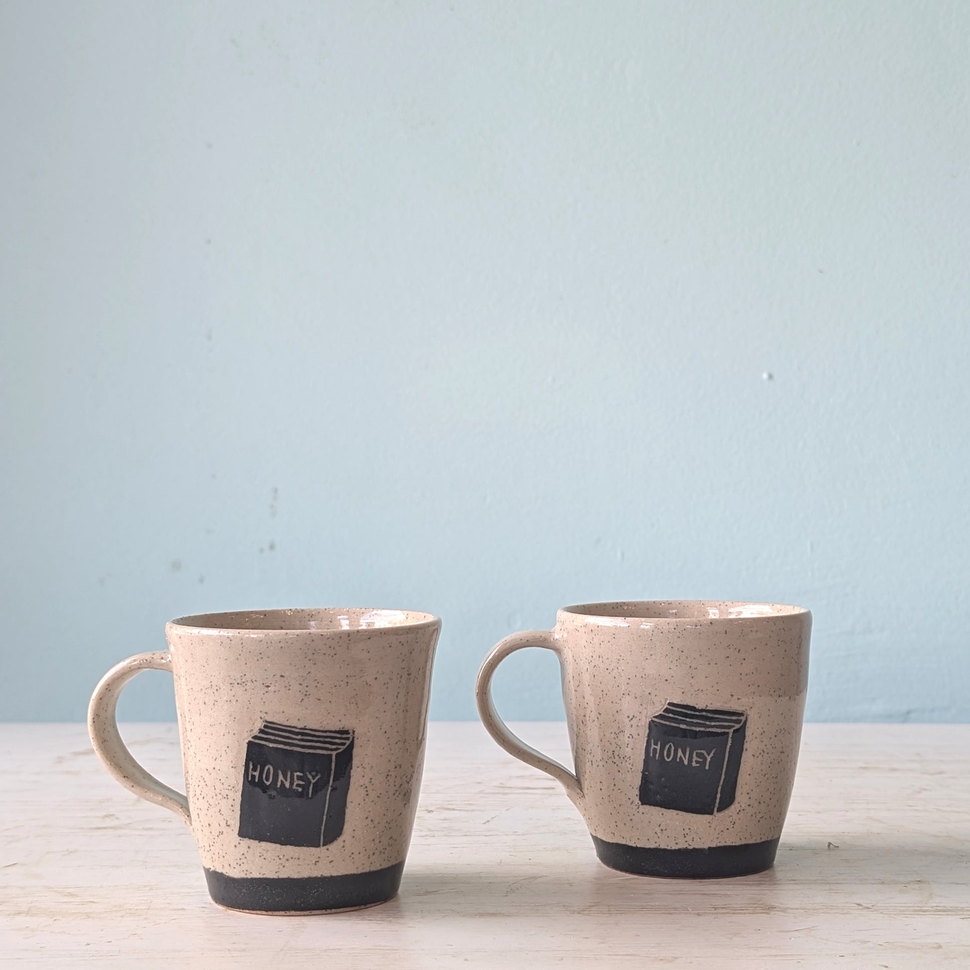 Two ceramic mugs with 'honey' design on a light blue background