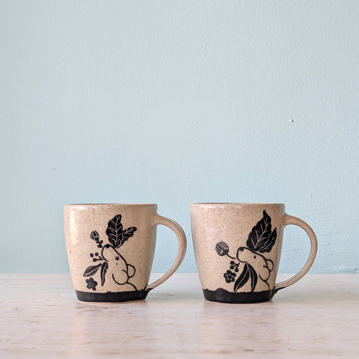 Two handmade ceramic mugs with a speckled beige glaze, decorated with black illustrations of a pika and leaves, sitting on a light wooden surface against a pale blue wall.
