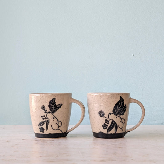 Two handmade ceramic mugs with a speckled beige glaze, decorated with black illustrations of a pika and leaves, sitting on a light wooden surface against a pale blue wall.