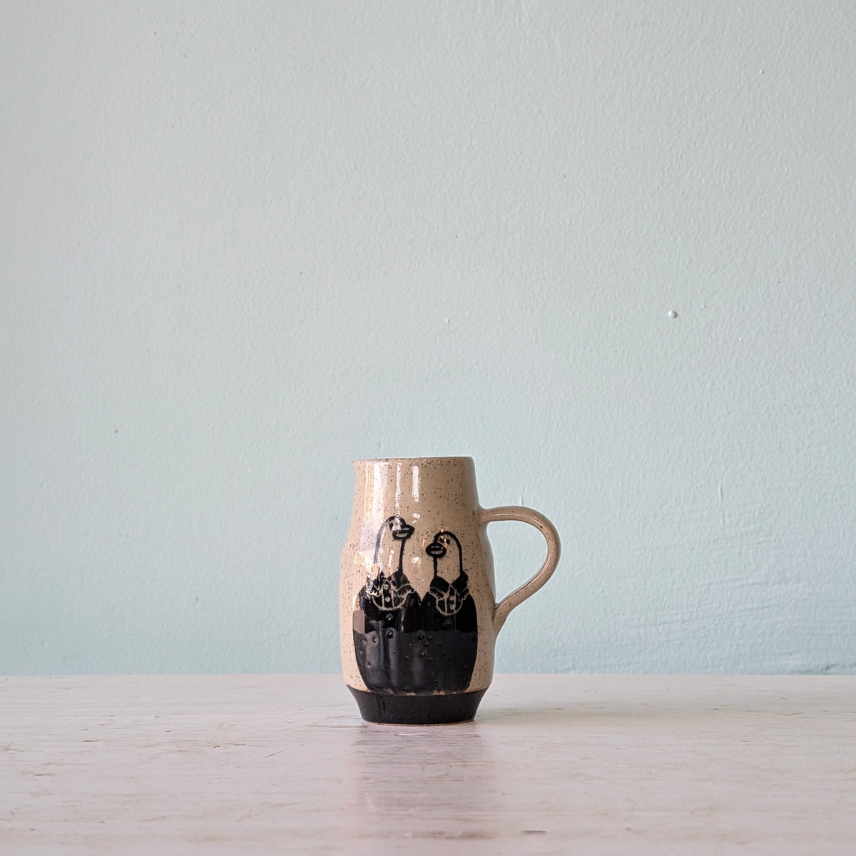 A handmade ceramic mug with a beige speckled glaze, black hand-painted goose figures, and a dark base, sitting on a light wooden surface against a pale blue wall.