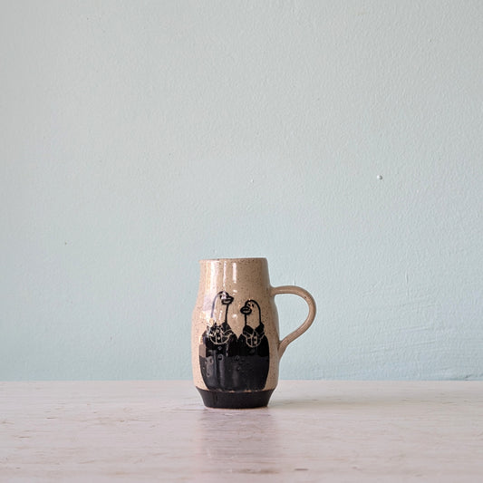 A handmade ceramic mug with a beige speckled glaze, black hand-painted goose figures, and a dark base, sitting on a light wooden surface against a pale blue wall.
