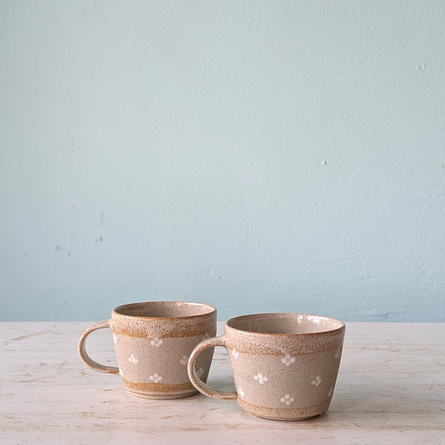 Two ceramic mugs with a speckled pattern on a light wooden surface and light blue background
