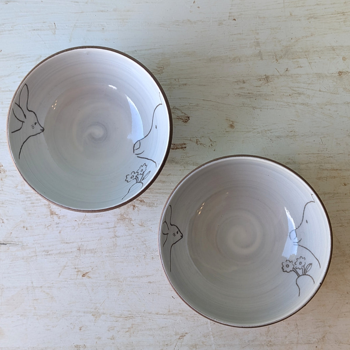 Two ceramic bowls with a white interior, hand-painted with simple black line drawings of animals and flowers, and finished with a dark brown rim, displayed on a light wood background.