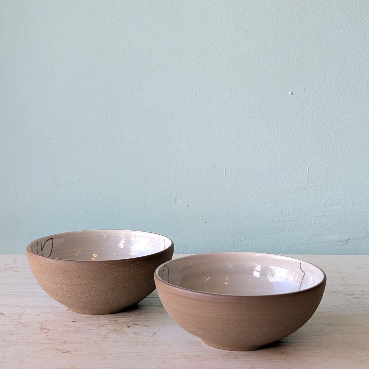 Two handmade ceramic bowls with a speckled beige exterior and a glossy off-white interior, displayed on a light wooden surface against a pale blue wall.
