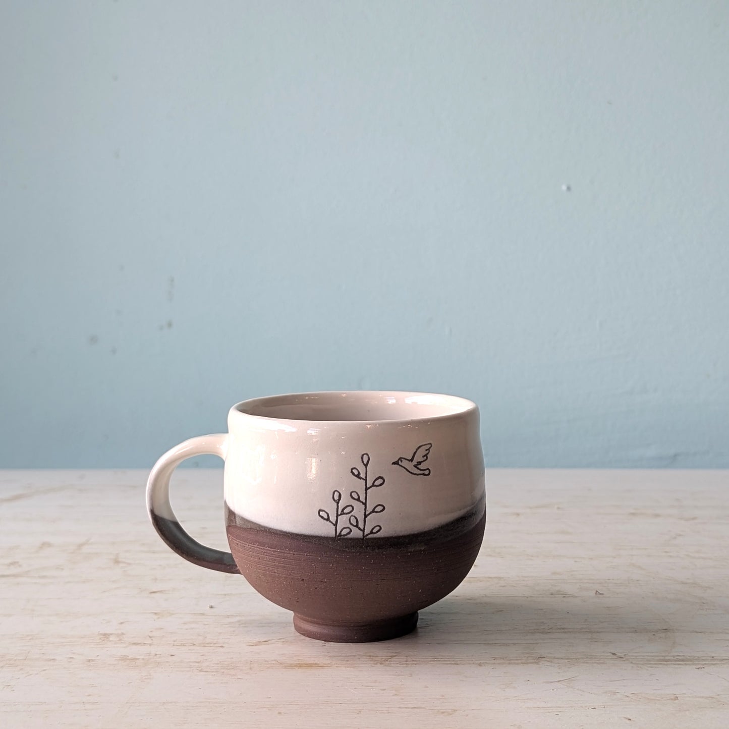 Ceramic mug with a brown bottom and white top on a light wooden surface with a light blue background