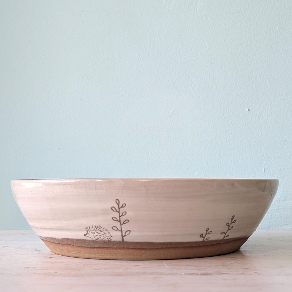 A wide, shallow ceramic bowl with a natural beige exterior and a brown glazed base, decorated with simple brown drawings of a hedgehog and plants. It is set against a light blue wall on a light wood surface.