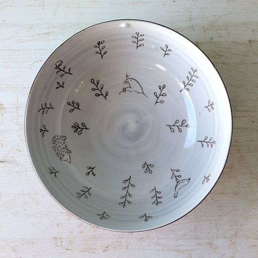 A round white ceramic bowl with a hand-painted brown design of small leaves, birds, and a hedgehog, photographed from directly above on a light wood background.