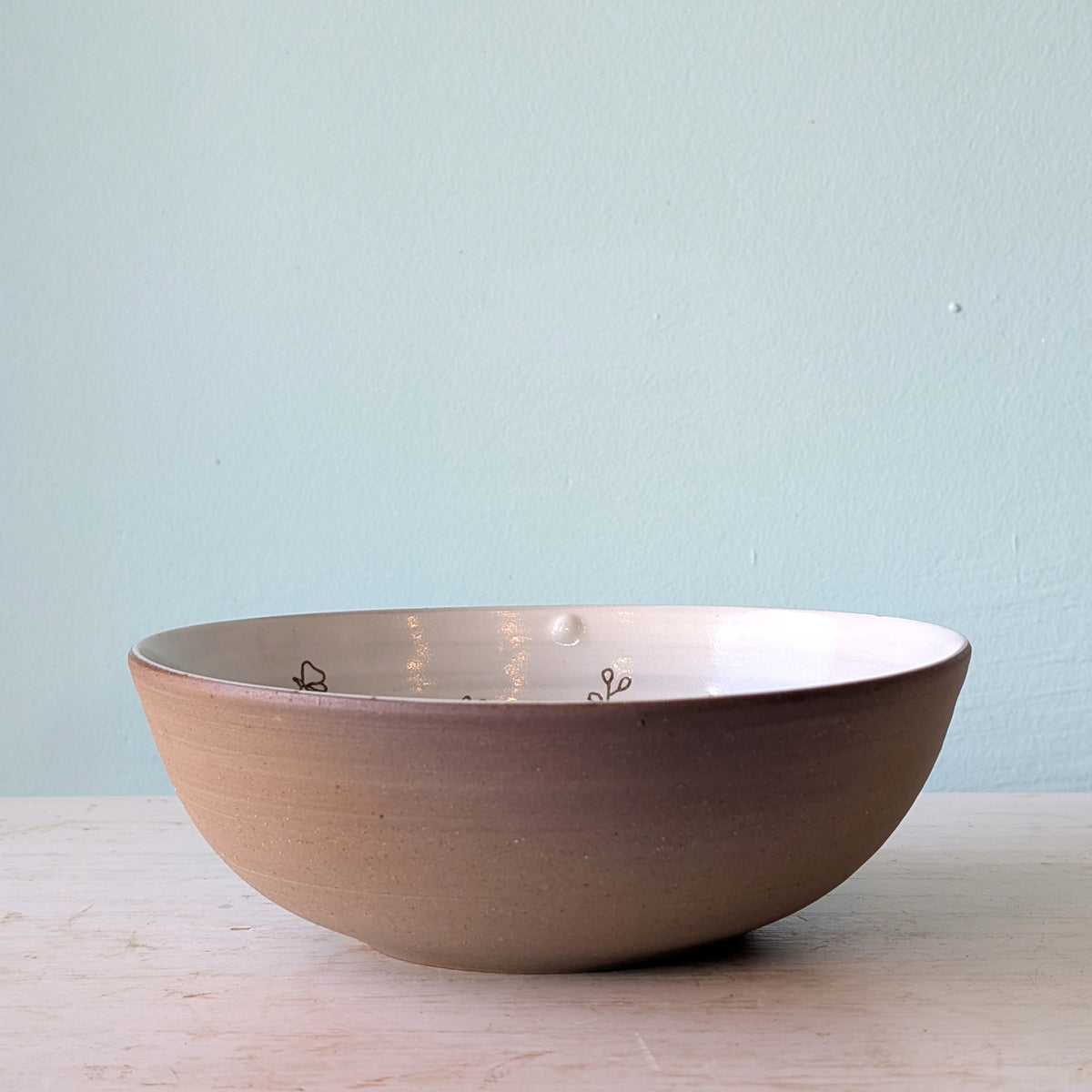 A handmade ceramic bowl with a textured, earthy brown exterior and a glossy, white glazed interior, sitting on a light wooden surface against a soft, pale blue wall.