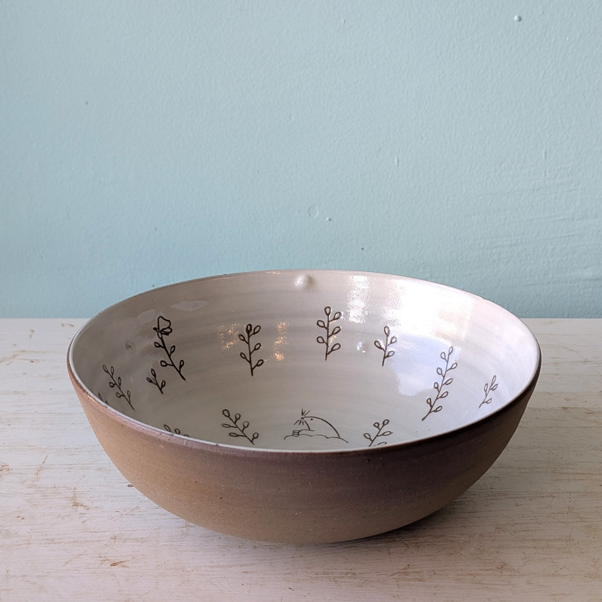 A handmade ceramic bowl with a matte brown exterior and a glossy white interior decorated with delicate hand-painted brown botanical patterns, resting on a light wooden surface against a pale blue wall.