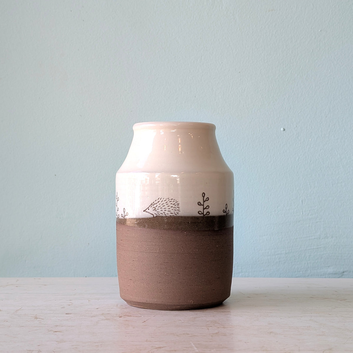 A handmade ceramic vase with a glossy white upper half, a textured brown lower half, and a hand-painted hedgehog design on the transition band, set against a light blue wall.