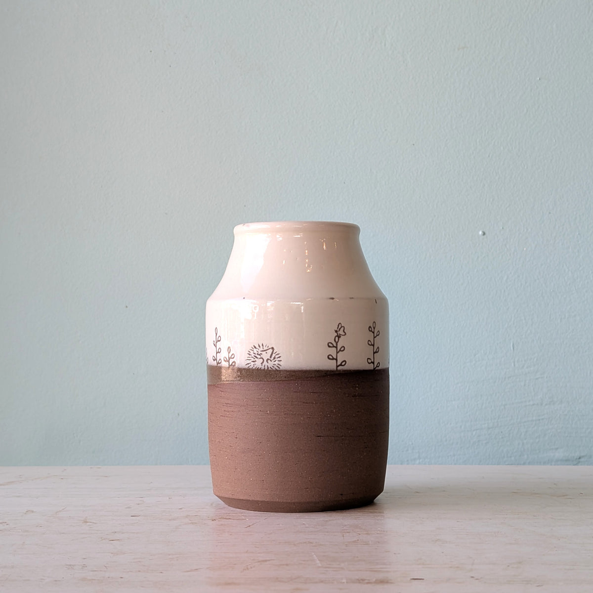 A handmade ceramic vase with a glossy white upper section, a textured brown middle band, and delicate hand-painted botanical motifs, displayed on a light wooden surface against a pale blue wall.