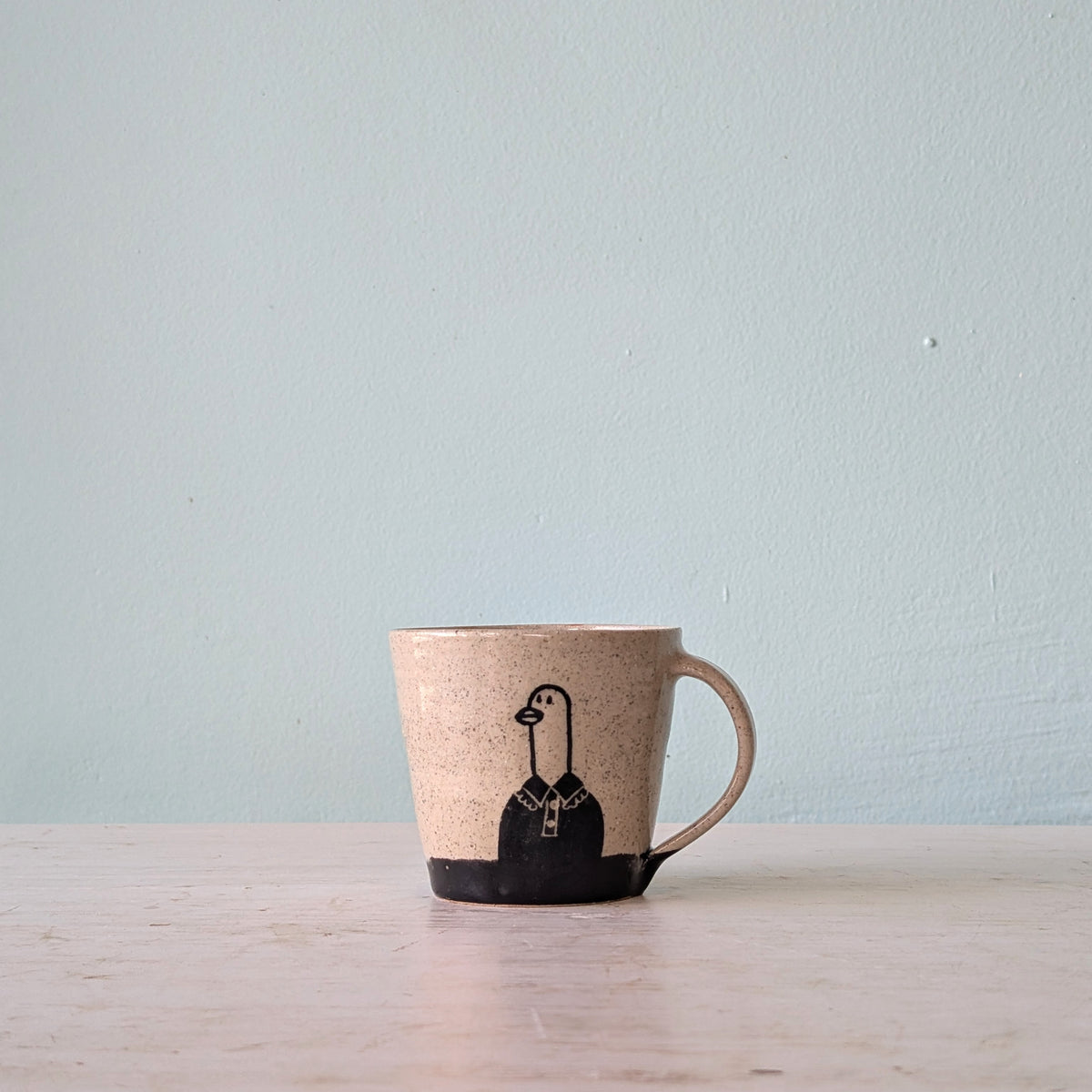 A handmade ceramic mug with a speckled beige body, a solid black base, and a black illustration of a goose wearing a shirt and tie, sitting on a light wooden surface against a pale blue wall.
