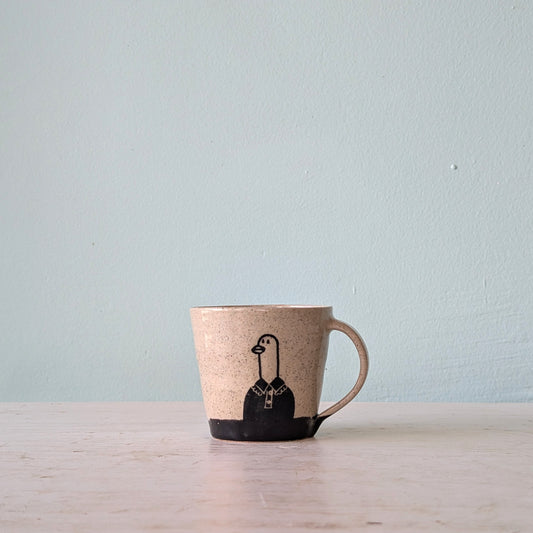 A handmade ceramic mug with a speckled beige body, a solid black base, and a black illustration of a goose wearing a shirt and tie, sitting on a light wooden surface against a pale blue wall.