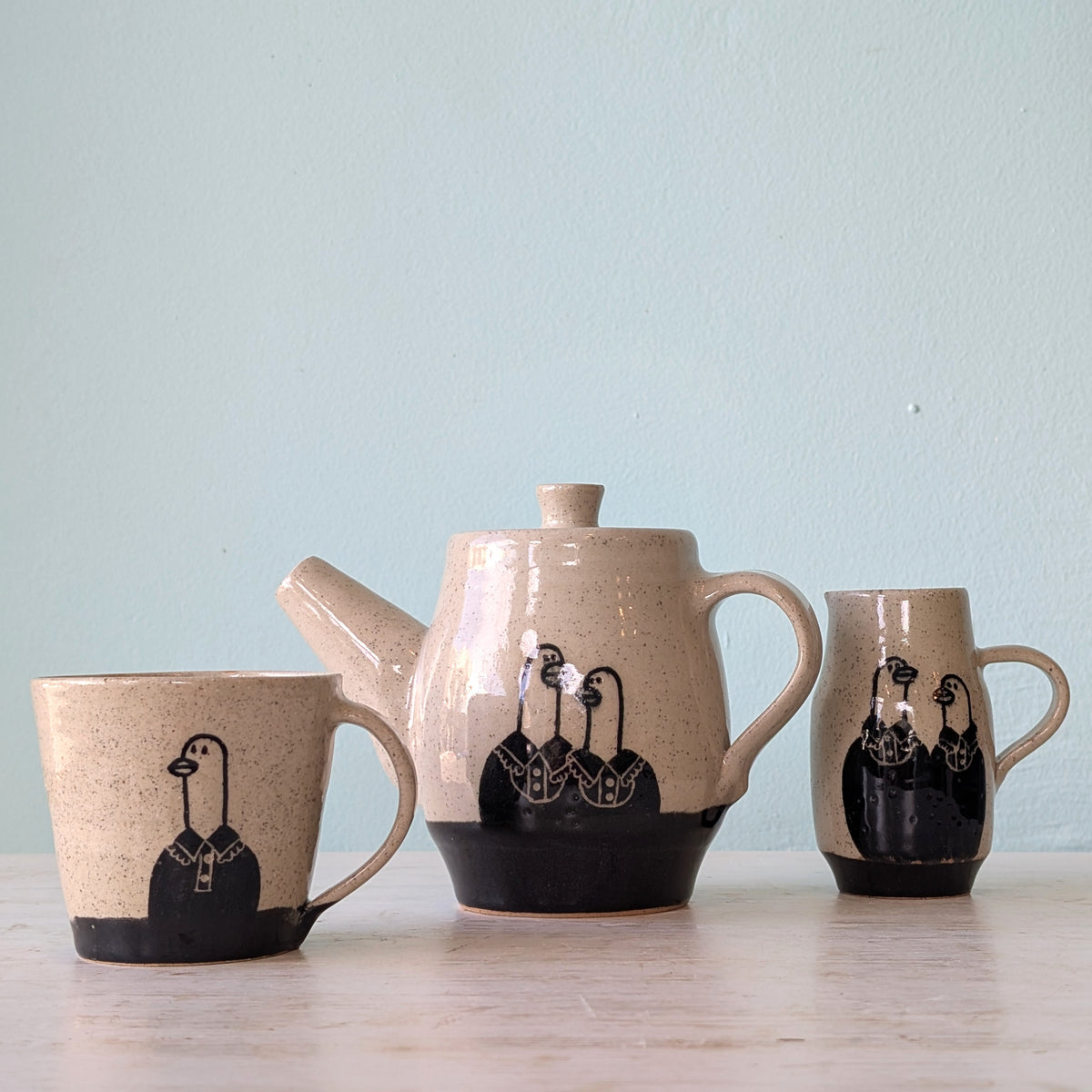 Three-piece ceramic tea set with a teapot, one cup and a milk jug, featuring a speckled beige glaze, black base, and stylized goose illustrations on a light wooden surface against a pale blue wall.
