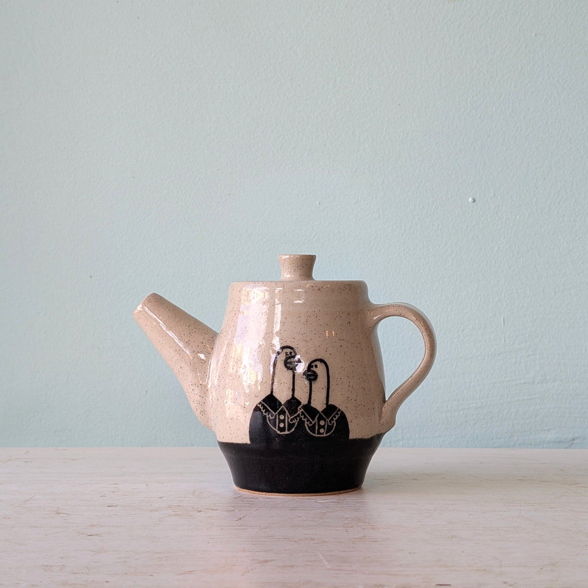 A handmade ceramic teapot with a speckled beige upper body, a black glazed base, and a black graphic of two geese, set on a light wooden surface against a pale blue wall.