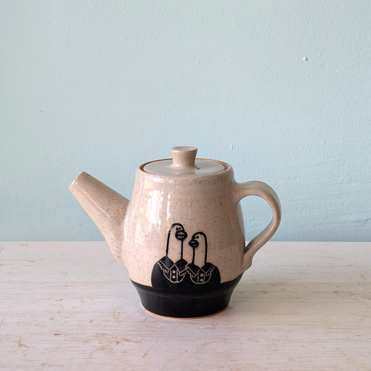 A handmade ceramic teapot with a speckled beige glaze, a black painted design of three stylized geese on the lower body, and a solid black foot ring, set on a light wooden surface against a pale blue wall.