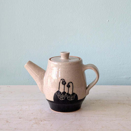 A handmade ceramic teapot with a speckled beige glaze, a black painted design of three stylized geese on the lower body, and a solid black foot ring, set on a light wooden surface against a pale blue wall.